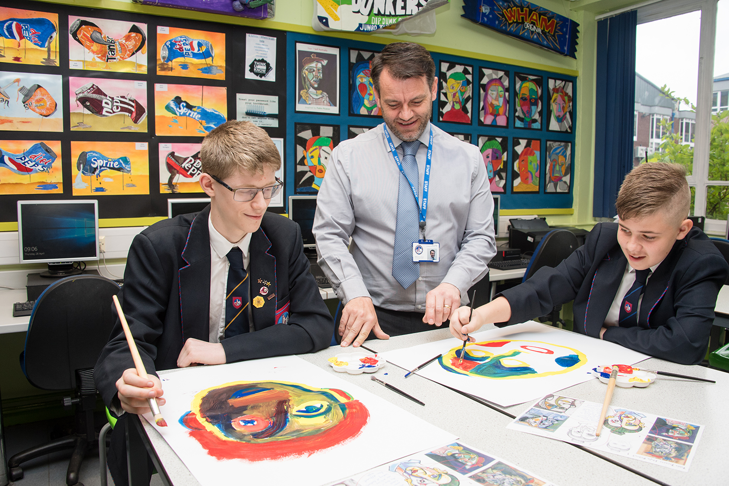 Two students painting in an art room with teacher standing in between them helping and smiling